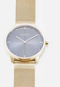 Reloj Calvin Klein con pulsera de malla dorada, caja redonda, esfera gris y marcadores de hora dorados. Presenta un diseño minimalista con un acabado elegante.