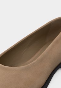 Κοντινό πλάνο ενός μπεζ σουέντ μπαλαρίνου μεσαίας μίας μπαλαρίνας που δείχνει την εσωτερική επένδυση και το μαύρο άκρο της σόλας πάνω σε λευκό φόντο.