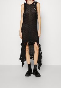 Svart stickad klänning med lång asymmetrisk fåll, utskurna sidor och fransdetaljer längs kanterna. Bärs med svarta ankelstövlar.