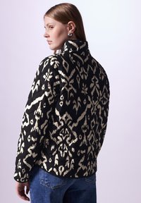 Gepolsterte Jacke im schwarz-weißen Ikat-Muster, mit hohem Kragen und langen Ärmeln. Glatte Textur, kombiniert mit Jeans.