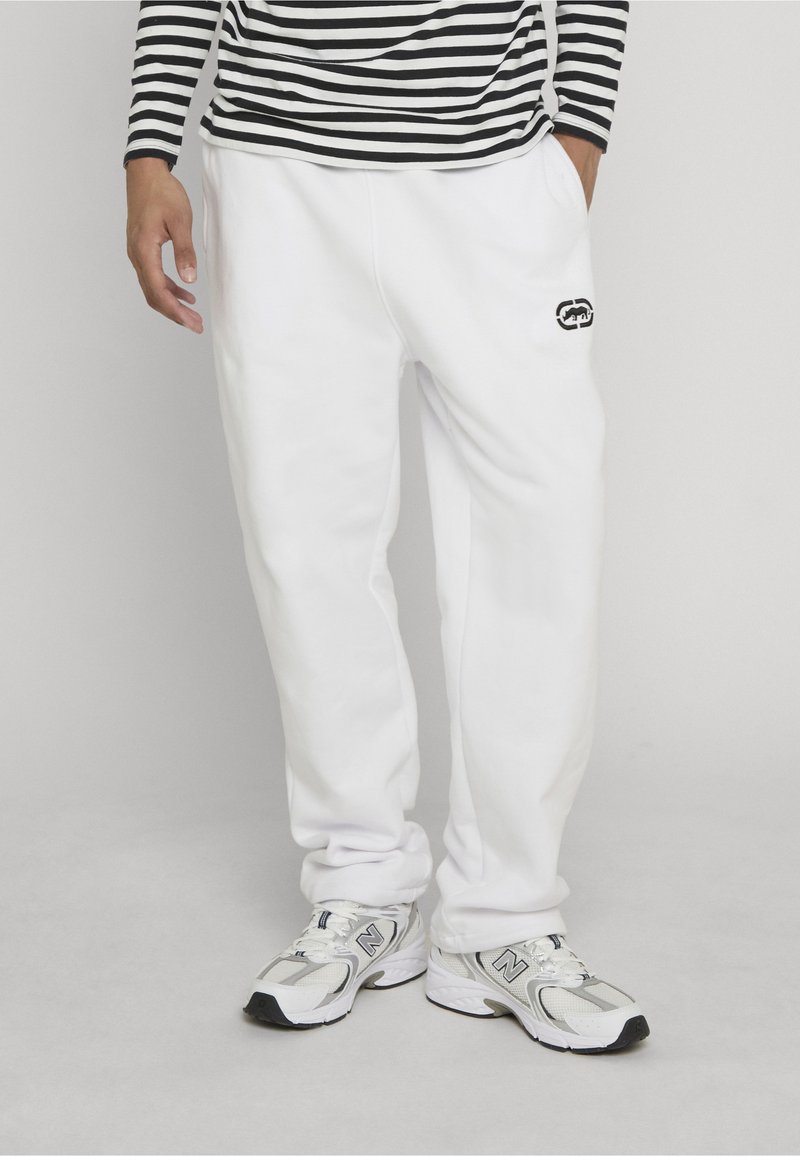 Personne portant un pantalon de survêtement blanc avec un petit logo noir sur la cuisse, un haut à manches longues rayé et des baskets blanches New Balance, debout sur un sol gris.