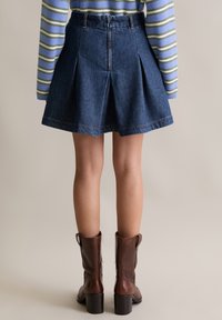 Gonna a pieghe in denim, lavaggio scuro, con dettagli di cucitura color sabbia. Abbinata con stivali al ginocchio marroni con tacco.