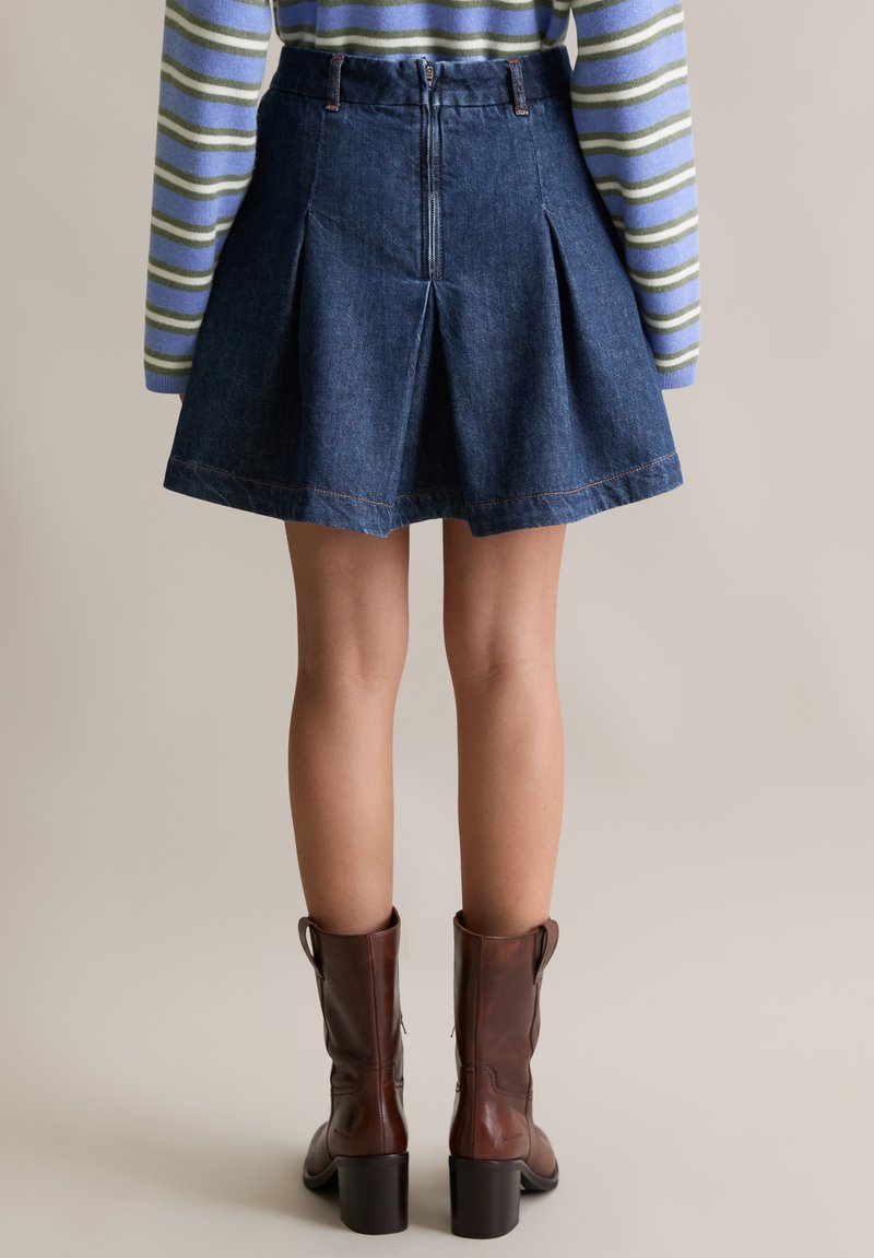 Gonna a pieghe in denim, lavaggio scuro, con dettagli di cucitura color sabbia. Abbinata con stivali al ginocchio marroni con tacco.