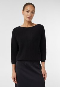 Schwarzer, gestrickter Pullover mit weitem Ausschnitt und Dreiviertelärmeln, kombiniert mit einem dunklen, leichten Rock. Geschmeidige Textur mit subtilen Details.