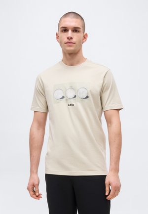 Lühikeste juustega mees kannab beeži T-särki, millel on kolm maapallo illustratsiooni ja allpool trükitud sõna "BOSS", mida kannab koos mustade pükstega.