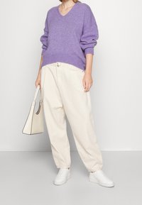 Maglione viola a scollo a V, in maglia morbida, abbinato a pantaloni beige larghi. Sneakers bianche, borsa rigida crema con finitura strutturata.