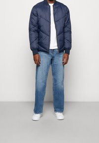 Marineblauwe pufferjack met chevronstiksels, ribgebreide manchetten en een ritssluiting. Gecombineerd met lichtblauwe spijkerbroek en witte sneakers.