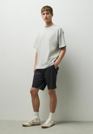 Junger Mann steht mit den Händen in den Taschen, trägt ein hellgraues, übergroßes T-Shirt, schwarze Shorts, weiße Socken und graue Turnschuhe vor einer schlichten Wand.