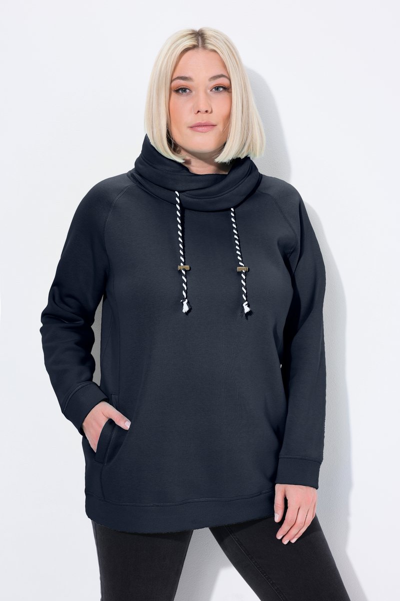 Blondínka v čiernej pulóvre s kapucňou s bielymi a čiernymi šnúrkami, jedna ruka v vrecku, na jednoliatom svetlom pozadí.