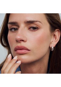 Silverfärgad ringformad örhänge med texturerad yta och stängning, bärs av en modell. Mjuka hudtoner och naturlig makeup, fingrar som försiktigt rör vid hakan.