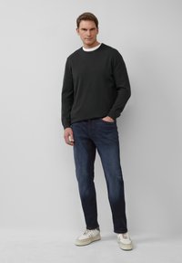 Donker sweatshirt met ronde hals, denim blauwe jeans en witte sneakers met crèmekleurige accenten; model staat tegen een effen lichte achtergrond.