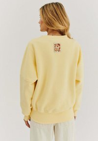 Žltá mikina s voľným strihom, rebrovanými manžetami a lemom. Na zadnej strane sa nachádza textúrovaná nášivka s ilustráciami jedla v hnedých odtieňoch.