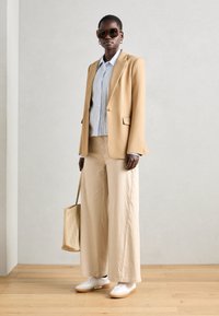 Blazer beige sur une chemise bleu clair, associé à un pantalon beige large et des baskets blanches. Accessoirisé avec de grandes lunettes de soleil et un sac fourre-tout.