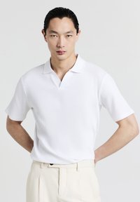 Polo shirt bianco a coste con scollatura a V, maniche corte e stile minimalista. Indossato con pantaloni di colore chiaro. Texture liscia.