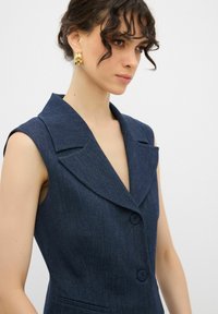 Ärmlös marinblå denimblazer med slagkrage, knäppning och sidofickor. Ytterligare synliga guldhoopörhängen.