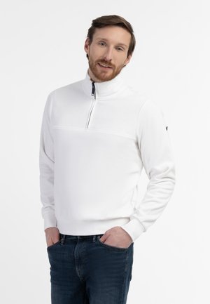 Mann mit hellbraunen Haaren und Bart, trägt weißen Quarter-Zip-Pullover und dunkelblaue Jeans, steht mit den Händen in den Taschen auf weißem Hintergrund.
