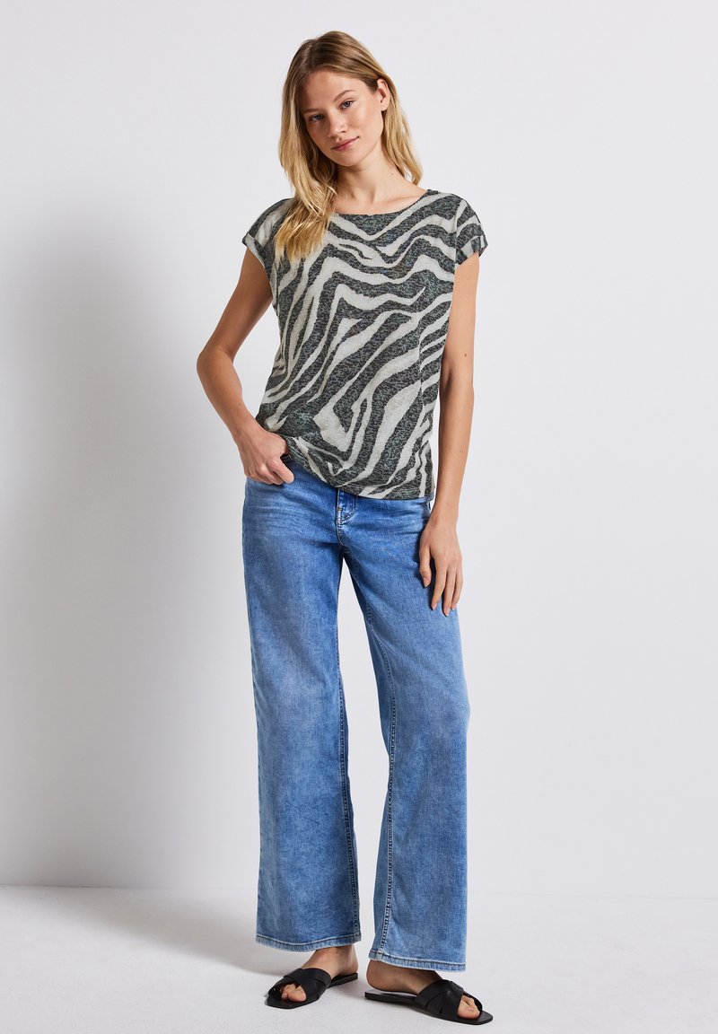 Top leggero a maniche corte con un motivo a righe zebrate verde e bianco, abbinato a jeans a gamba larga blu chiaro e sandali neri scivolati.