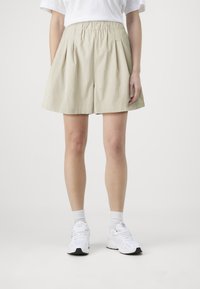 Pantaloni corti plissettati beige realizzati in tessuto leggero, dotati di una vita elastica e una vestibilità morbida, abbinati a sneakers bianche.