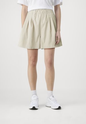 Beige plissert shorts laget av lett materiale, med elastisk liv og en avslappet passform, sammen med hvite sneakers.