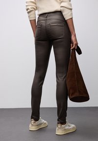 Jeans ajustados de mezclilla elástica de color marrón oscuro, con una textura elegante, bolsillos traseros y un bolso de ante marrón a juego. Suéter en tonos neutros y zapatillas visibles.