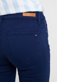 Jeans de mezclilla azul oscuro con un parche de cuero etiquetado "Tommy Hilfiger" y un pequeño logo en rojo, blanco y azul en el bolsillo trasero.