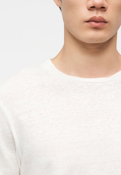 Κοντινό πλάνο του κάτω μέρους του προσώπου και του λαιμού ενός ατόμου που φοράει λευκό, υφασμάτινο μπλουζάκι με στρογγυλή λαιμόκοψη σε ανοιχτό φόντο.