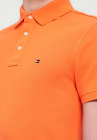 Πορτοκαλί πόλο με γιακά, κατασκευασμένο από υφή ύφασμα και μικρό κεντημένο λογότυπο στο στήθος σε κόκκινο, άσπρο και μπλε.