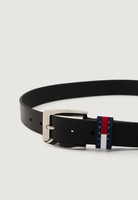 Cinturón de cuero negro con una hebilla de metal plateado y una etiqueta de marca en azul, rojo y blanco que lleva el texto "TOMMY JEANS".
