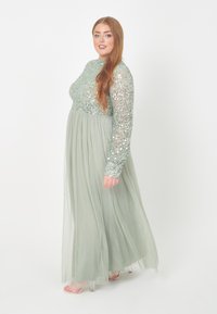 Langes, hellgrünes Kleid mit einem mit Pailletten verzierten Oberteil, langen Ärmeln und einem fließenden Rock. Klare Riemchensandalen ergänzen das Outfit.