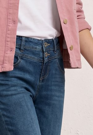 Person iført blå jeans med høj talje og dobbelt knappelukning, hvid skjorte stukket ned i, og pink jakke med guldknapper.
