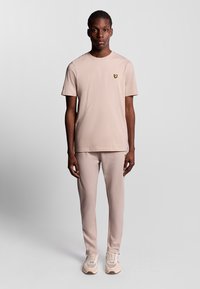 Lys beige kortærmet t-shirt med et lille gult logo, parret med matchende slim-fit bukser og beige sneakers, der har en glat struktur.