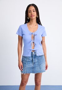 Top scurt cu maneci scurte, de culoare mov deschis, cu decolteu în V și detalii cu fundă în față. Asortat cu o fustă mini din denim cu margini desfăcute.