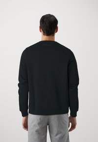 Felpa nera con maniche lunghe, collo rotondo e polsini a coste. Presenta un logo in rilievo discreto sul retro. Abbinata a pantaloni della tuta grigi.
