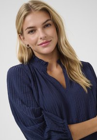 Blusa blu navy texture con scollo a V e colletto arricciato. Maniche lunghe con polsini arricciati. Il modello ha i capelli biondi lunghi e lisci.
