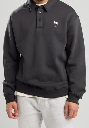 Uomo con felpa polo nera a maniche lunghe con piccolo logo bianco della bandiera di NYC sul petto e pantaloni della tuta grigio chiaro, in piedi davanti a uno sfondo semplice.