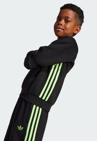 Schwarzer Trainingsanzug mit drei vertikalen neongrünen Streifen an den Ärmeln, gerippten Bündchen und Taillenbund. Mit dem Adidas-Logo auf der Brust.