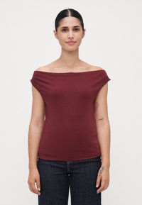 Mujer con cabello oscuro, vistiendo una blusa marón sin mangas y jeans oscuros, de pie frente a un fondo blanco liso, mostrando tatuajes en su brazo izquierdo.