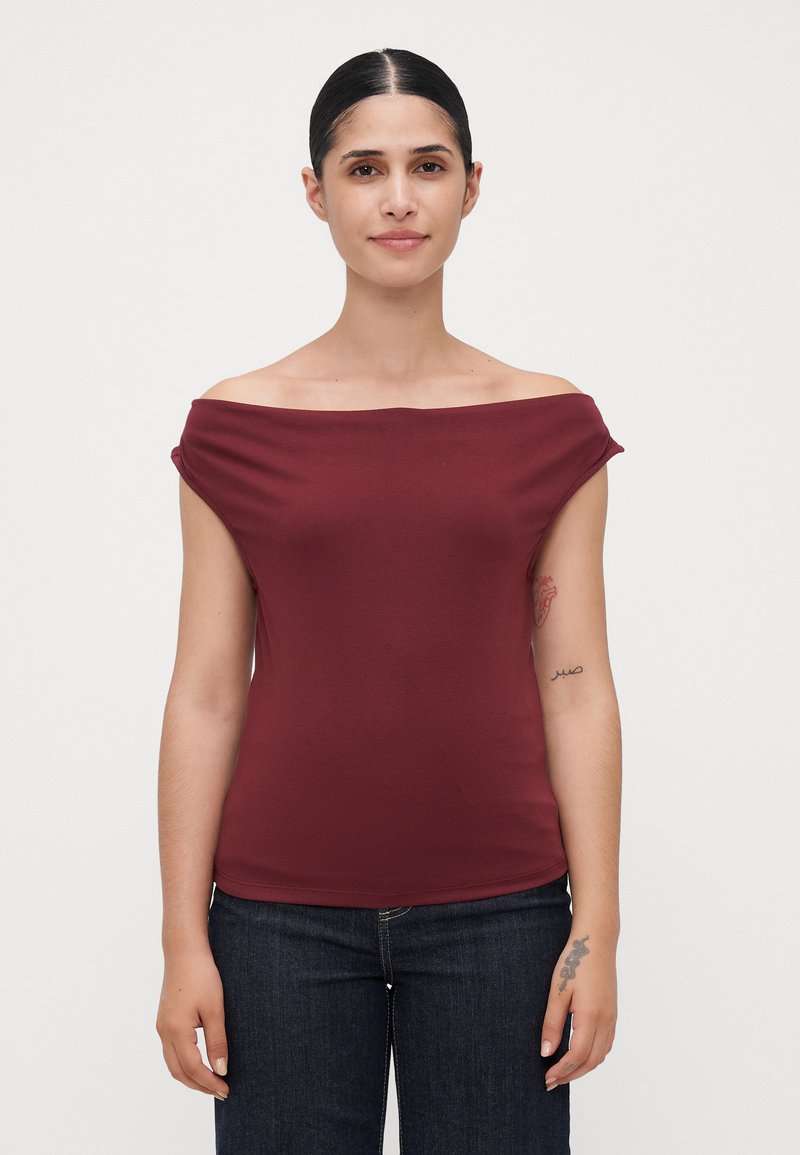 Mujer con cabello oscuro, vistiendo una blusa marón sin mangas y jeans oscuros, de pie frente a un fondo blanco liso, mostrando tatuajes en su brazo izquierdo.