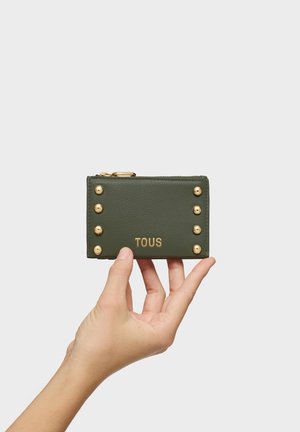 Hand holding a green TOUS leather wallet with gold studs and a gold zipper against a plain white background.