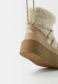 Beige højtopstøvle med fluffy shearling-overdel, flad brun gummisål, dekorative metalnitter og mønstrede snørebånd.