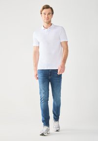 Fehér póló, kék szűk farmert és szürke tornacipőt visel. A póló rövid ujjú és galléros, míg a farmer enyhén kopott denim.