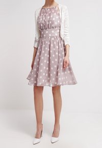 Světle růžové šaty s puntíky, se střiženým pasem a rozšířenou sukní, doplněné bílým krajkovým kardiganem a bílými lodičkami.