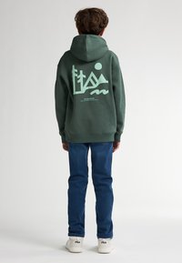 Groene hoodie met een grote mintgroene grafische afbeelding op de achterkant, gecombineerd met blauwe jeans en witte sneakers, die een casual outfit laat zien.