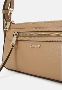 Saco crossbody em pele bege com detalhes em dourado, apresentando um bolso com fecho e logótipo empotrado. Textura suave com realces em castanho contrastante.