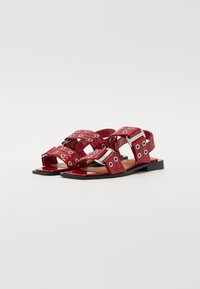 Red patent leather sandals with multiple straps, circular cutouts, and silver hardware accents. Flat sole, open-toe design.