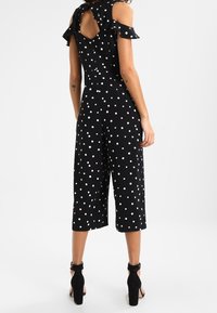 Combinaison noire à pois avec épaules dénudées, manches volantes, détail en forme de clé à l'arrière et design de jambes larges raccourcies. Portée avec des talons noirs.