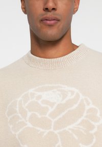 Beige pulóver, fehér virágmintával, kerek, bordázott nyakkivágással, puha tapintással és testhez simuló fazonnal.