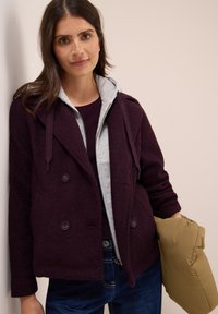 Veste burgonde à double boutonnage avec une finition texturée, présentant une capuche grise en dessous, associée à une blouse beige à manches bouffantes et un jean marine.