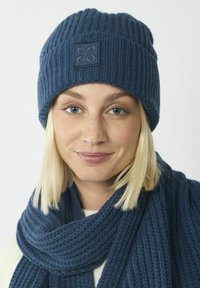 Marine gebreide muts met ribbelstructuur en geborduurd vierkant patch. Aangevuld met een bijpassende dikke sjaal, beide in vergelijkbaar materiaal.
