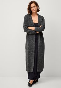Langer grauer Cardigan mit strukturiertem Strick, offener Vorderseite und gerippten Bündchen, über einem schwarzen Kleid getragen, kombiniert mit flachen schwarzen Schuhen.